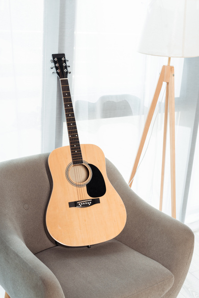 Guitarra acústica na poltrona perto da lâmpada de assoalho perto da janela na sala de estar
 - Foto, Imagem