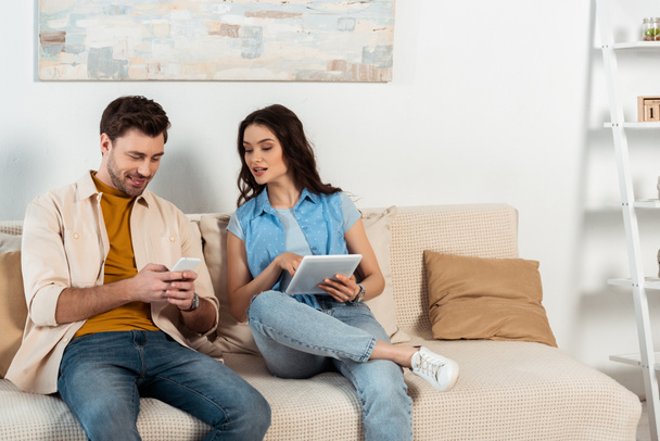 Smiling man using smartphone near girlfriend with digital tablet at home  - Photo, Image