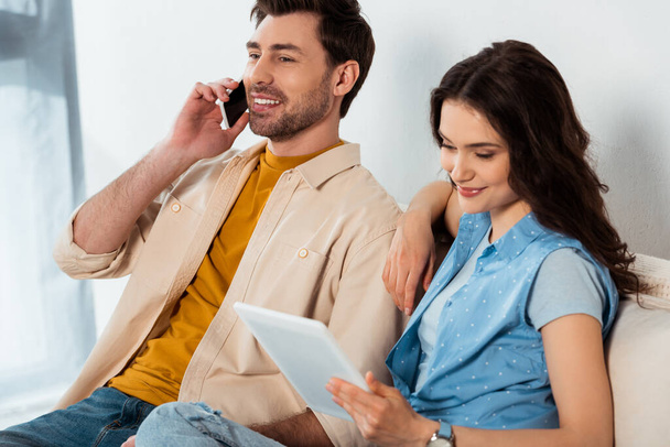 Selective focus of smiling man talking on smartphone near girlfriend using digital tablet at home  - Foto, Bild