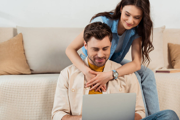 Smiling girl embracing boyfriend using laptop at home  - Фото, зображення