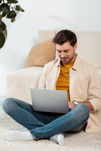 Smiling freelancer using laptop while sitting on floor in living room - Фото, изображение