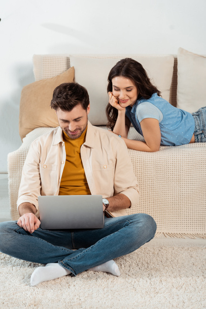 Smiling woman lying on couch near boyfriend using laptop on floor  - Photo, Image