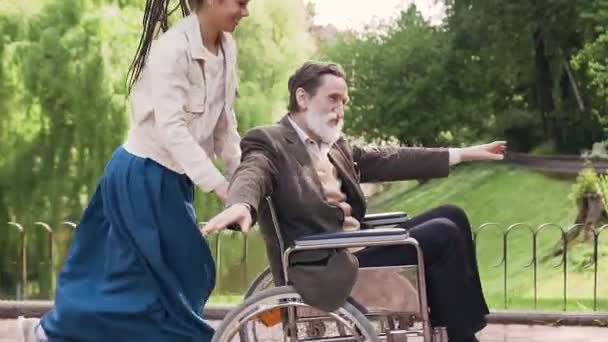 Cheerful good-looking carefree modern young girl with dreadlocks and elderly disables-man in wheelchair having fun together while walking in green park - Materiał filmowy, wideo