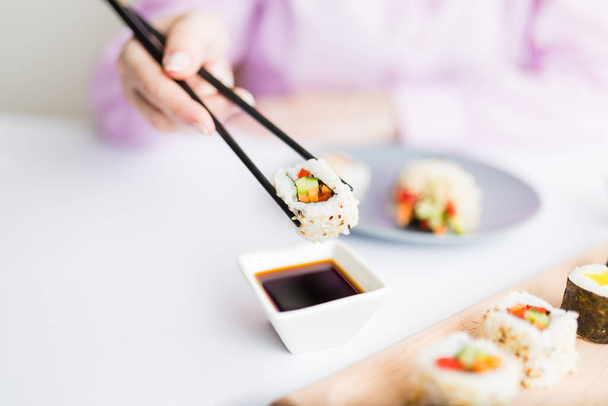Close up of woman holding sushi roll with chopsticks above bowl full of soy sauce. Healthy traditional japanese meal concept. - Фото, зображення