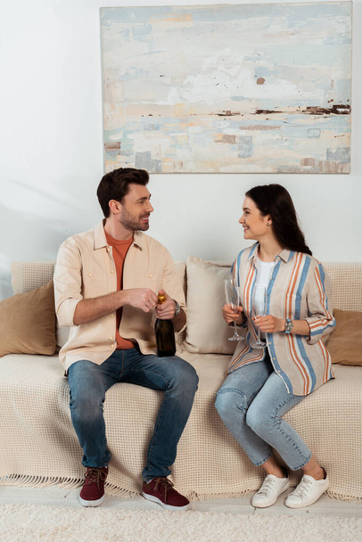 Smiling woman holding glasses and looking at boyfriend opening bottle of champagne at home  - Photo, image
