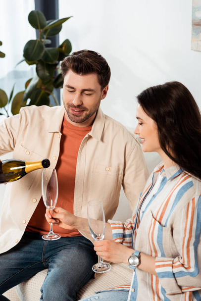 Smiling woman holding glass while boyfriend pouring champagne on couch  - Foto, afbeelding