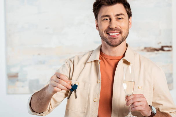 Smiling man looking at camera while holding keys of new house and glass of champagne  - 写真・画像