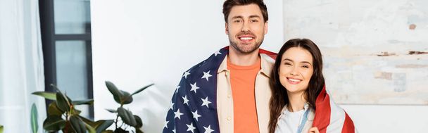 Panoramablick auf lächelndes Paar, das sich in amerikanische Flagge hüllt und in die Kamera blickt  - Foto, Bild