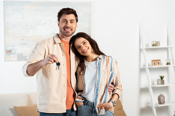 Young couple smiling at camera while holding keys of new house in living room - Zdjęcie, obraz