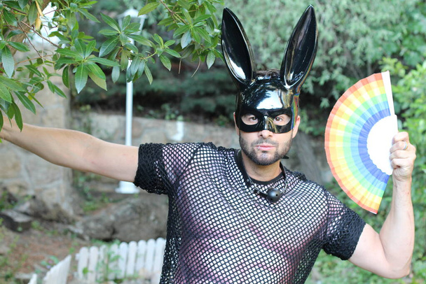 close-up portret van knappe jonge man in gay trots kostuum met konijn masker en lgbt gekleurde handheld ventilator op straat - Foto, afbeelding