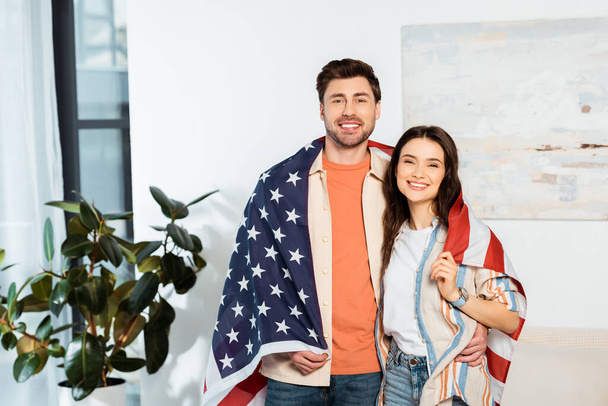Schöner Mann umarmt lächelnde Freundin, während er sich in amerikanische Flagge hüllt  - Foto, Bild