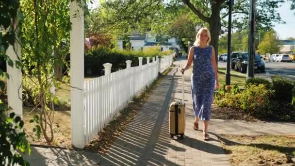 A woman with a travel bag on wheels is walking along the sidewalk. A typical American town. Steadicam shot - Filmmaterial, Video