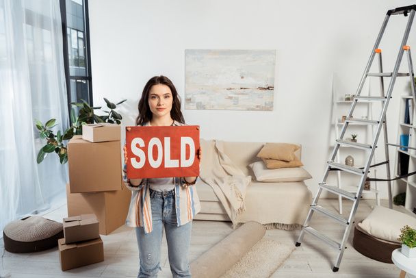 Atractiva chica sosteniendo placa con letras vendidas cerca de cajas de cartón y escalera en la sala de estar
 - Foto, Imagen