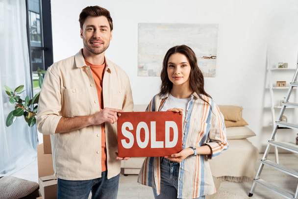 Smiling man holding nameplate with sold lettering near girlfriend in living room - Photo, Image