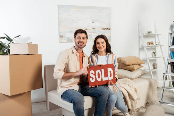 Cheerful couple looking at camera while holding nameplate with sold lettering near carton boxes at home  - Photo, Image
