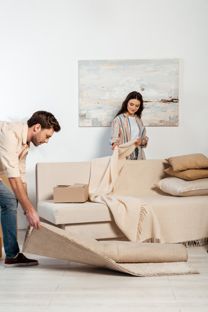 Selective focus of woman holding plaid near couch while boyfriend turning off carpet in living room - Φωτογραφία, εικόνα
