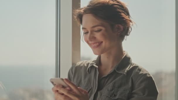 Pleased young pretty woman using smartphone while standing near the window at home - Footage, Video