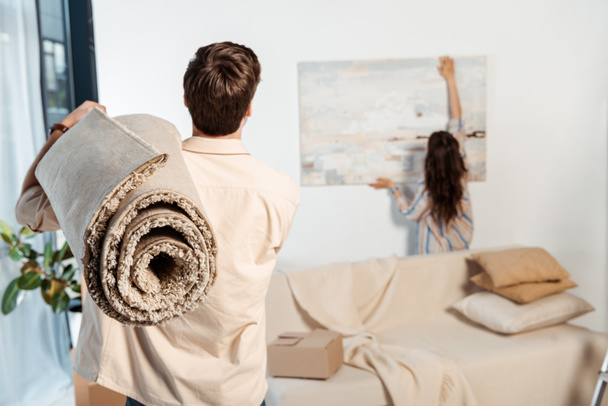 Visão traseira do homem segurando tapete enquanto a namorada tomando pintura da parede durante o movimento
  - Foto, Imagem