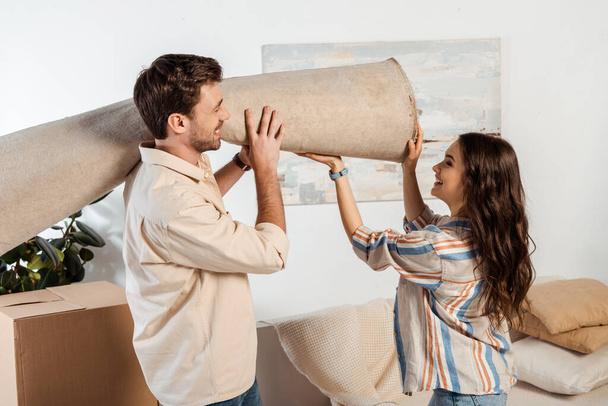 Vue latérale du couple souriant tenant le tapis près de la boîte en carton dans le salon - Photo, image