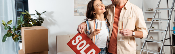 Panoramic shot of smiling woman holding glass of champagne near boyfriend with sold lettering on nameplate in living room - Photo, Image