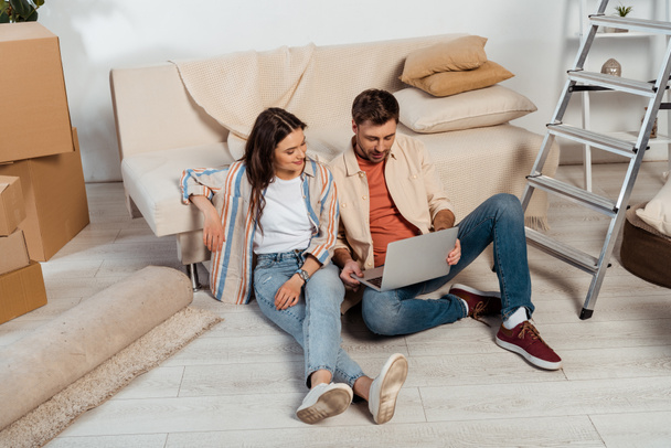 Smiling woman sitting near boyfriend using laptop in new house  - Photo, Image