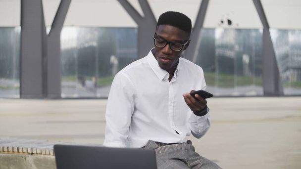 Attractive African Businessman with smart phone looking at notebook and recording voice message in park - Photo, image