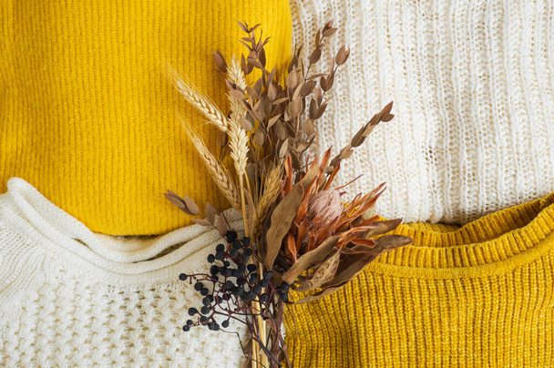 Pile of knitted clothes yellow white color with dried flower bouquet. Autumn winter concept. - Foto, Bild