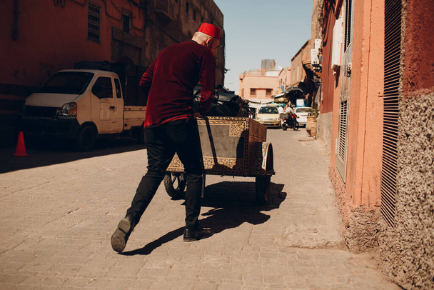 Cansado porteiro com carrinho pesado e bagagem turística andando na rua para o hotel em Marraquexe, Marrocos
. - Foto, Imagem