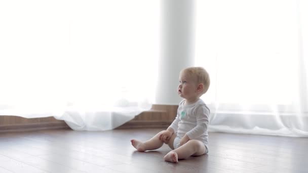 Kleines Baby im weißen Body sitzt auf einem warmen Holzboden. - Filmmaterial, Video