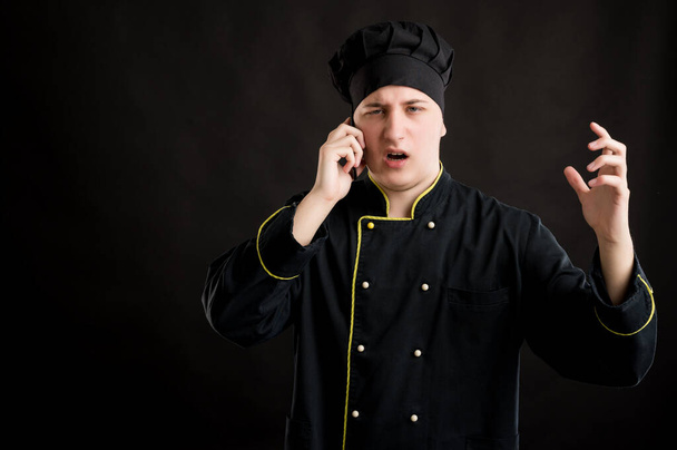 Portret van jonge man gekleed in een zwart chef-kok pak met bruin haar, misnoegd gezicht en gebaren en praten op mobiele telefoon, poseren op een zwarte geïsoleerde achtergrond. - Foto, afbeelding