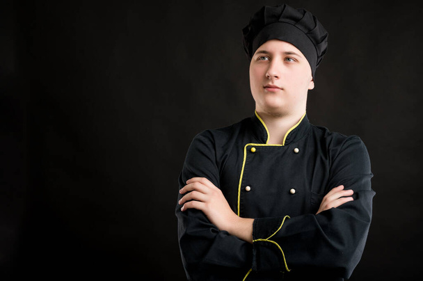Portrét mladého muže oblečeného v černém kuchařském obleku, který vypadá sebejistě hrdina-shot pózuje na černém izolovaném pozadí s kopií prostoru reklamní oblasti - Fotografie, Obrázek