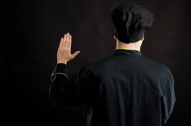 Portrait of young male dressed in a black chef suit showing oath from behind posing on a black isolated background with copy space advertising area - Foto, Imagen
