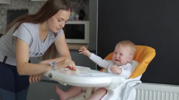 Mom feeds the baby in a feeding chair. The child cries and does not want to eat - Imágenes, Vídeo