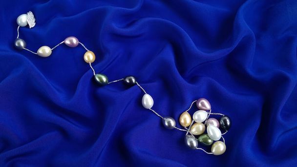 A multicolored pearl necklace is beautifully laid out on dark blue silk. Large natural iridescent  luxury pearls ideally harmonize with  the blue  navy velvety background. Shot from above. - Photo, Image