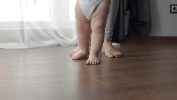 Ensimmäiset askeleet. Pieni lapsi oppii kävelemään äitinsä kädestä. Valkoihoinen lapsi ottaa ensimmäiset askeleensa puulämmitteisellä lämpimällä lattialla - Materiaali, video