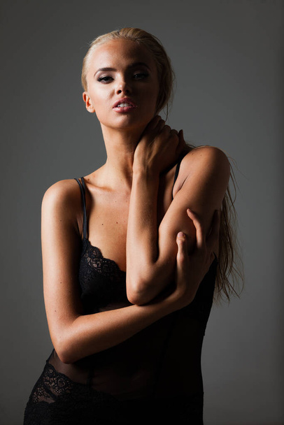 Portrait of blond young woman with beautiful makeup and hairstyle in a black lace bodysuit stands on gray background. Fashion model poses in elegant clothes in studio - Φωτογραφία, εικόνα