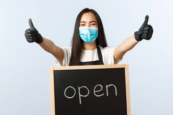 Small business, covid-19 pandemic, preventing virus and employees concept. Upbeat asian female store worker, cafe staff in medical mask invite visitors, show thumb-up and we are open sign - Photo, image