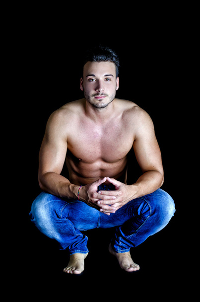 Shirtless muscular young man in jeans sitting on dark background - Foto, Bild