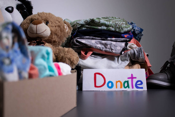 Donate sign handwritten with multicoloured letters. A box with clothes and a pile of clothes nearby on a grey table.clothes donation concept. copy space - Photo, Image