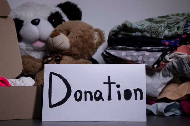 Spendenschild handgeschrieben mit schwarzen Buchstaben. Eine Schachtel mit Kleidung und ein Stapel Kleider in der Nähe auf einem grauen Tisch. Kopierraum - Foto, Bild