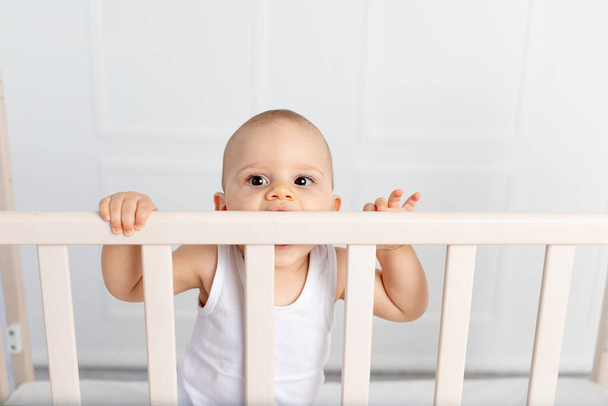portrait of a baby boy 8 months old standing in a crib in a children's room in white clothes and nibbling on the bed, baby's morning, children's goods concept - Photo, Image