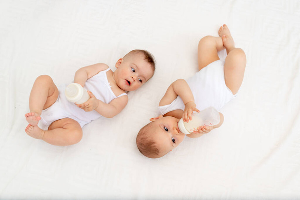 twee kinderen een jongen en een tweeling van 8 maanden drinken melk uit een fles op het bed in de kinderkamer, voeden de baby, baby food concept, bovenaanzicht, plaats voor tekst - Foto, afbeelding