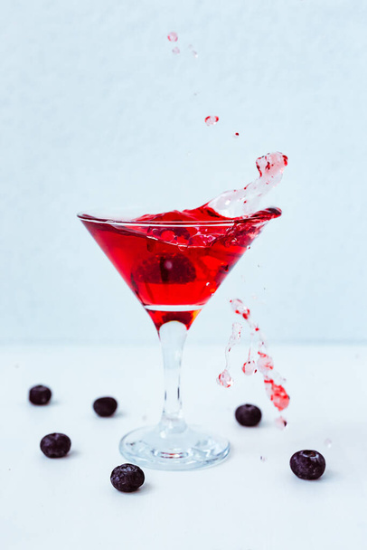 A photo of a red cocktail splash, martini glass, blueberries around, light blue background, natural light vertical photo - Valokuva, kuva