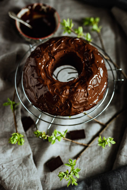 Chocolate bundt cake spring still life. Cake covered with chocolate icing. Young green leaves around, a bowl with melted frosting. Grey and beige linen table cloth - Fotografie, Obrázek