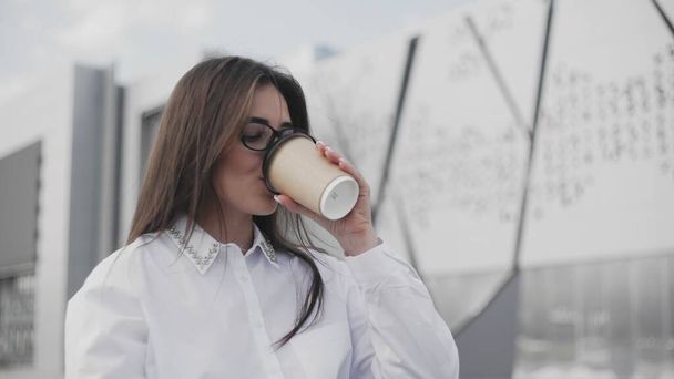 Mulher de negócios bonita em óculos conversando no smartphone e bebendo café enquanto sentada no parque perto de seu escritório
 - Foto, Imagem
