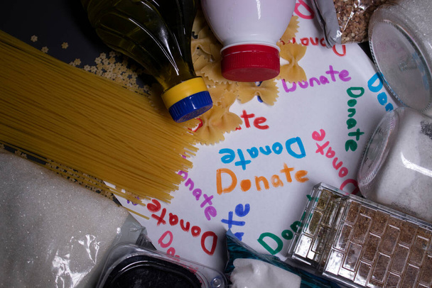 "Lahjoita "merkki käsin kirjoitettu monivärisiä kirjaimia. Kasa ruokatarvikkeita harmaalla pöydällä. Ruoan luovutuksen käsite. Coronavirus-pandemian tausta. kopioi tila - Valokuva, kuva