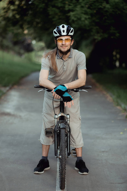 Portret van de mens in een gele zonnebril en helm staand met de fiets op de weg en kijkend in de camera - Foto, afbeelding