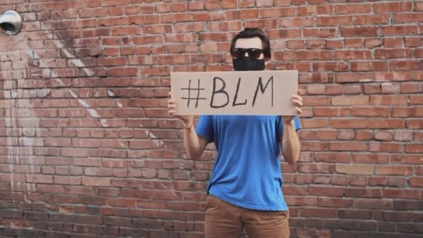 O homem caucasiano na máscara está contra a parede vermelha com o cartaz de papelão em mãos com a hashtag de inscrição - BLM. Protesto único
. - Filmagem, Vídeo