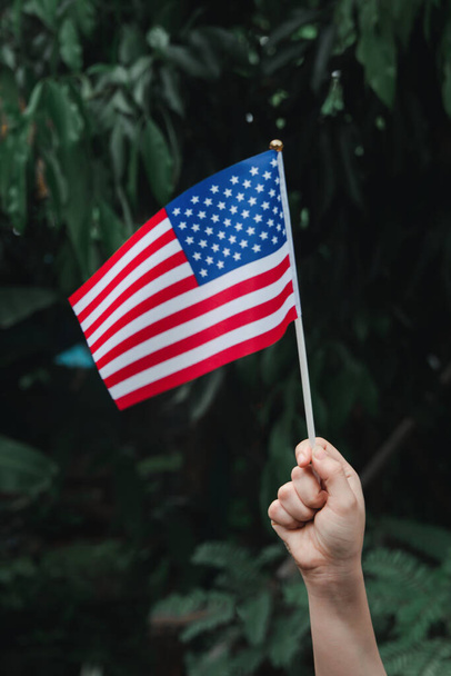 Γυναίκα χέρι κρατώντας σημαία των ΗΠΑ στο πράσινο δάσος. 4η Ιουλίου Ημέρα Αμερικανικής Ανεξαρτησίας - Φωτογραφία, εικόνα