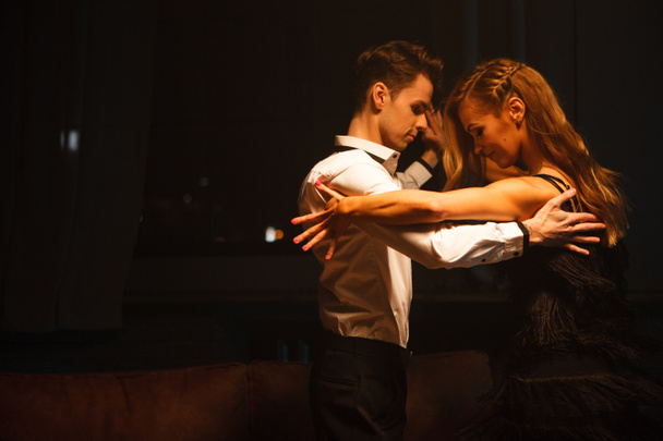 Dance and love concept. Young couple in elegant evening dresses posing in the room filled with dramatic light. Two dancers man and woman holding each other in passionate pose - Photo, Image